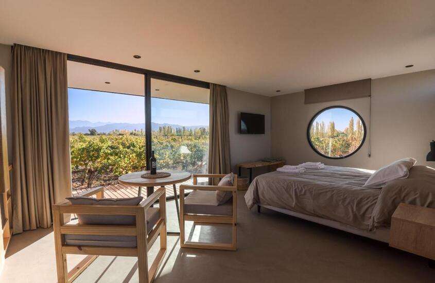 durante o dia, cama de casal, tv, cadeiras e mesa em suíte de hotel vinícola em Mendoza com vista para vinhedos