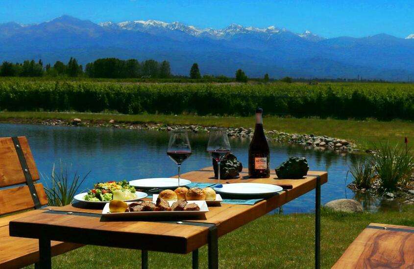 durante o dia, tábua de frios, taças de vidro e garrafa de vinho servidos em mesa de madeira ao ar livre com vista para lago e montanhas