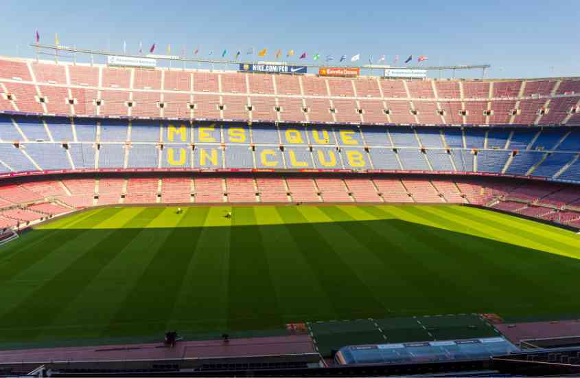 durante dia, sol iluminando arquibancada vermelha e azul e campo de futebol camp nou, ponto turístico de barcelona
