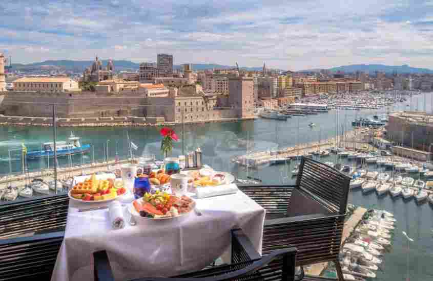 durante o dia, mesa forrada com toalha branca, onde há diversas frutas, queijos e frios, e cadeira marrom em varanda ao ar livre com vista para o mar