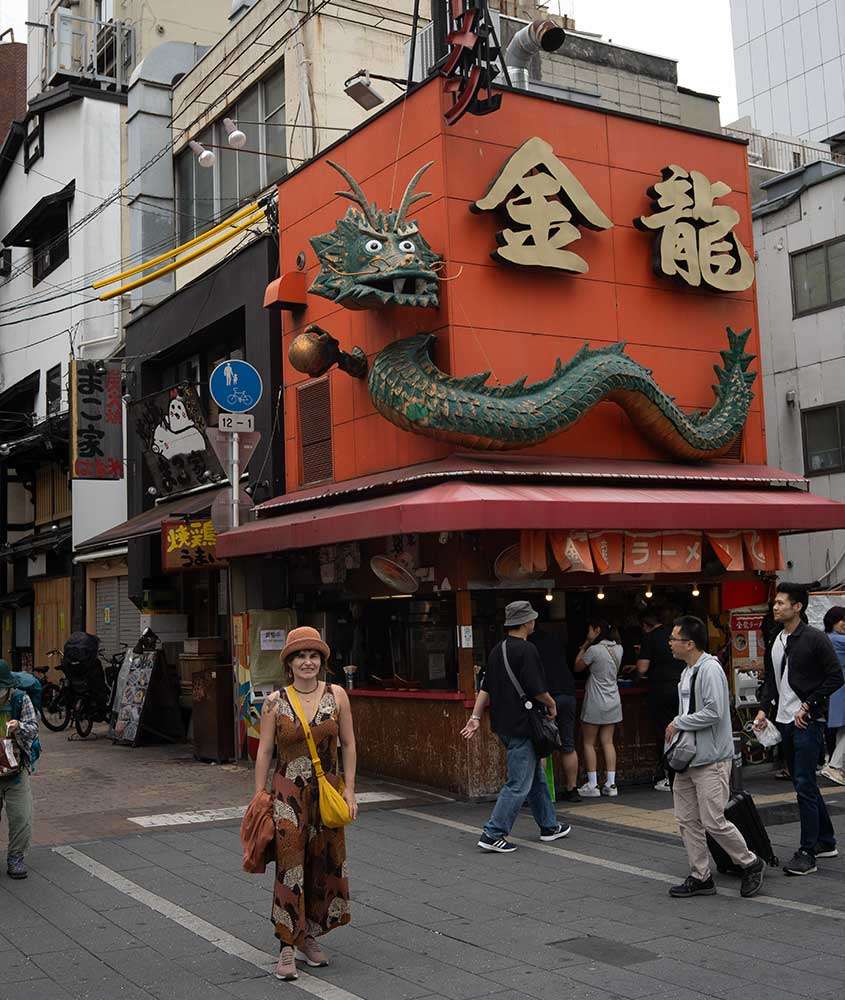 durante o dia, uma mulher em pé, sorri pra uma foto em rua de Osaka. Ao redor, há várias pessoas caminhando