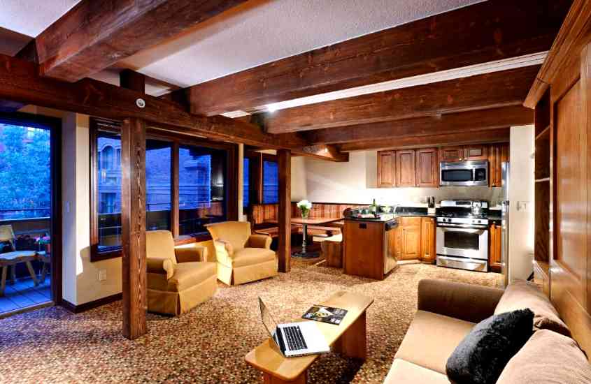 sofá, notebook em cima de mesa, poltronas, armário de madeira e fogão em suíte de hotel em aspen com varanda e vista para árvores