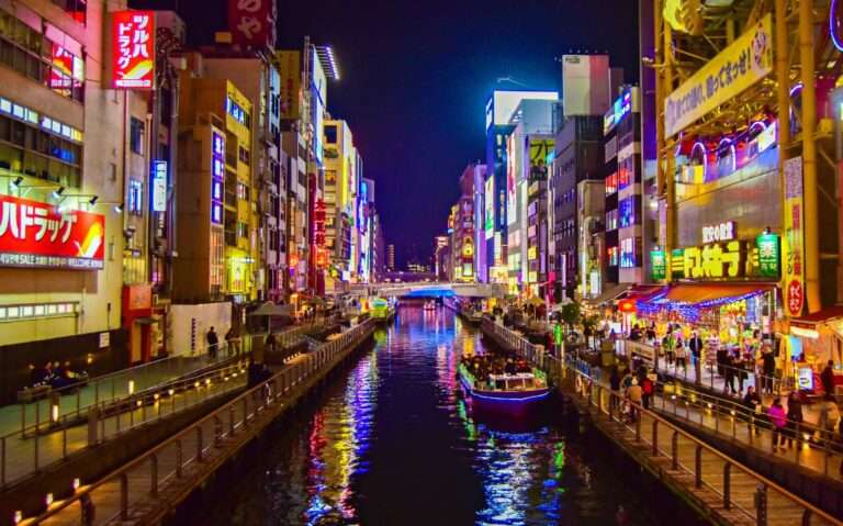 durante a noite, pessoas em barco iluminado passando em rio cercado de prédios com letreiros coloridos e frases em japonês em osaka japão