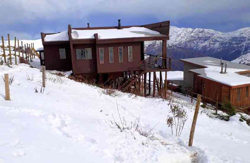 durante o dia, casa marrom, onde funciona um dos hotéis no valle nevado, com vista para as montanhas nevadas