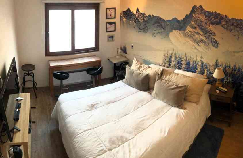 televisão, cama de casal, cadeiras e mesa, em suíte com pintura de montanhas nevadas