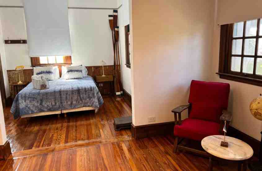 cama de casal, poltrona vermelha e pequena mesa redonda em suíte de hotel com chão de madeira