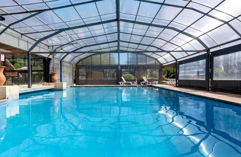 durante o dia, espreguiçadeiras em frente a grande piscina retangular em área de lazer de hotel coberta com teto de vidro