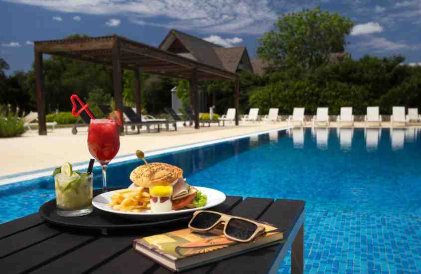 durante o dia, agenda, óculos de sol, prato com sanduíche e dois copos de suco em cima de mesa de madeira em frente a grande piscina ao ar livre