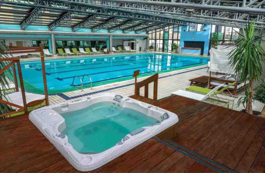 durante o dia, jacuzzi e grande piscina retangular em área de lazer de hotel coberta, com janelas de vidro