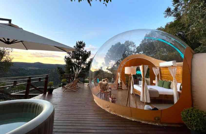 durante amanhecer, cama de casal, abajur, cadeira e telescópio em bolha de vidro climatizada, em frente a varanda com banheira e vista para as montanhas