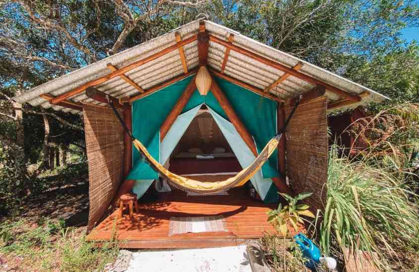 durante o dia, cama de casal, banco de madeira e rede de descanso em tenda de madeira em jardim de camping de luxo no brasil