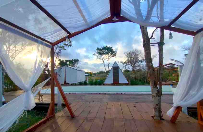 durante entardecer, tenda de madeira em formato triangular em frente a piscina de glamping no brasil