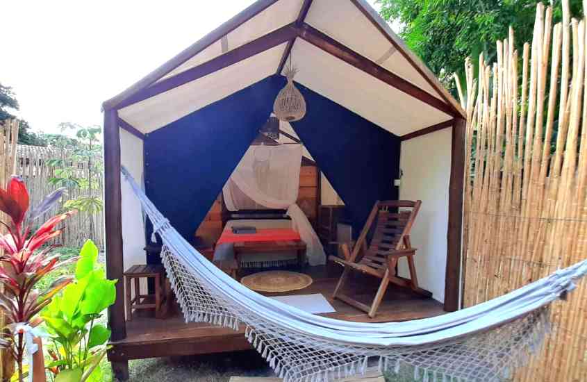 durante o dia, cama de casal, cadeira de madeira e rede de descanso em chalé de madeira em jardim de um dos hotéis glamping no brasil