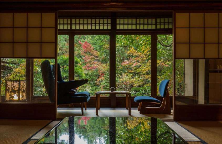 durante o dia, mesa quadrada e duas poltronas azuis em suíte de um dos ryokans em kyoto com vista para jardim cheio de árvores