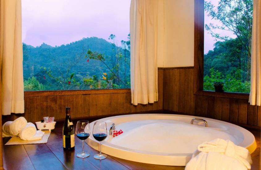 durante o dia, duas taças de vidro e garrafa de vinho em cima de deck de banheira de hidromassagem redonda, em suíte com vista para montanhas e árvores