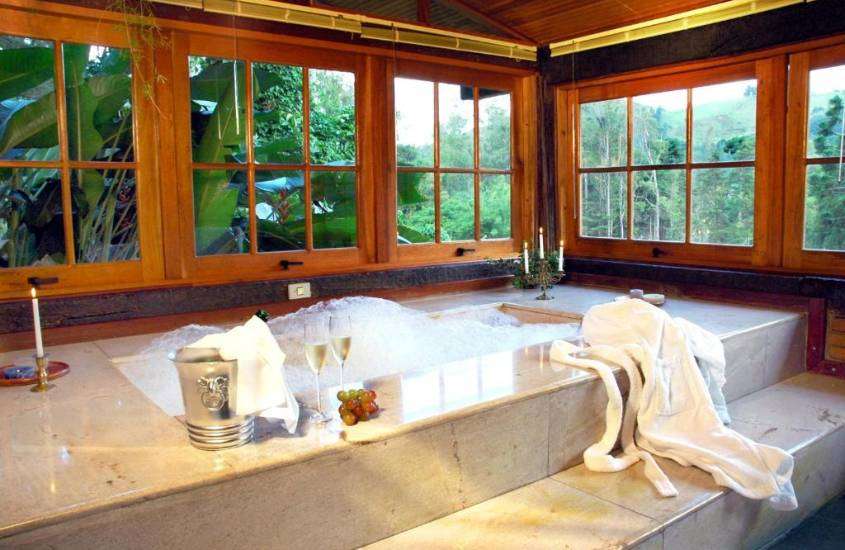 durante o dia, velas, taças de champagne e uvas em borda de banheira retangular de mármore, em banheiro com vista para montanhas e árvores