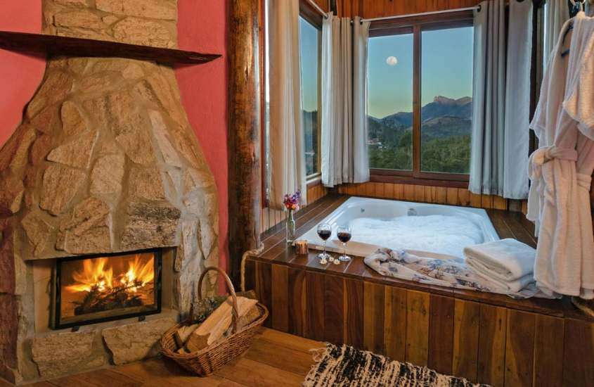 durante entardecer, lareira e taças de vinho em cima de borda de banheira quadrada em suite com vista para as montanhas