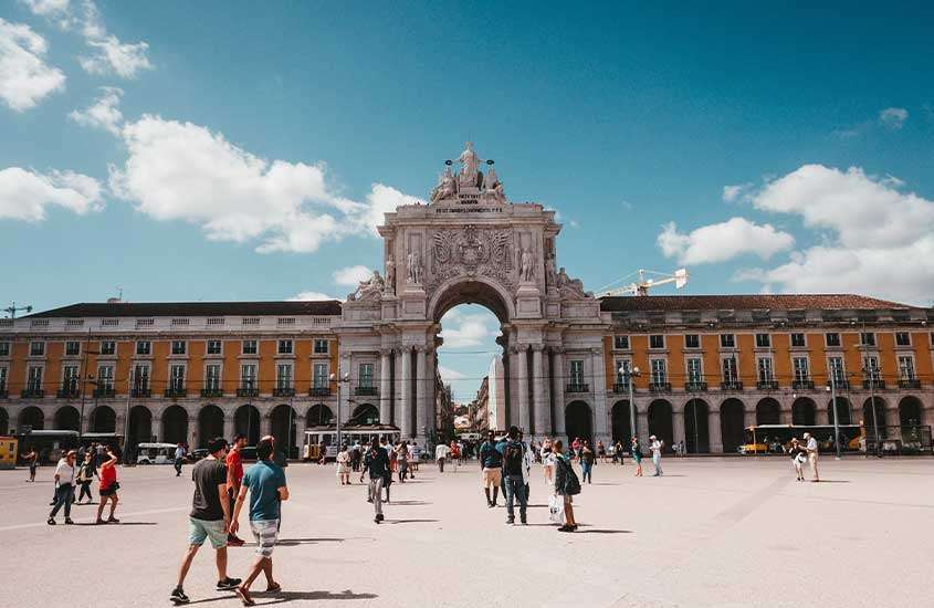 Durante o dia, pessoas caminhando na Praça do Comércio, um dos principais pontos turísticos de Lisboa, com um grande arco no fundo e uma estátua de um rei no centro da praça