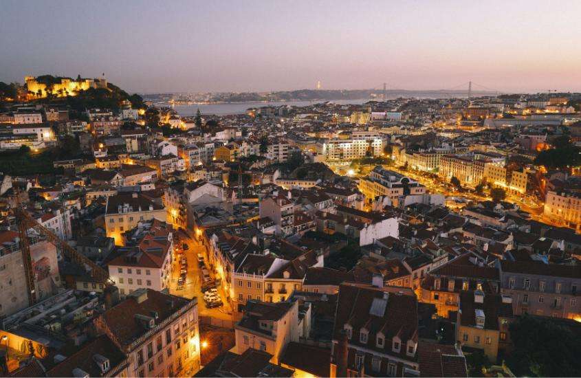 durante entardecer, vista aérea de casas, prédios e carros e, ao fundo mar, em bairro alto, um dos melhores bairros de Lisboa