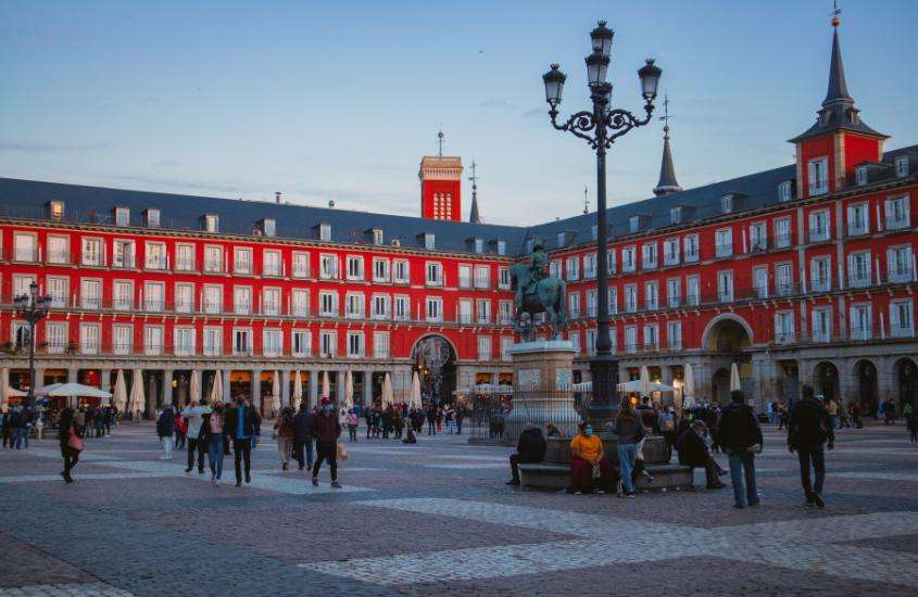 Durante o dia, várias pessoas caminhando e desfrutando da Plaza Mayor em Madrid, cercada por prédios históricos e coloridos