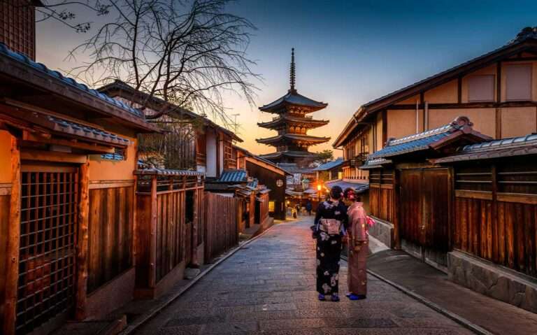 Duas gueixas, com suas tradicionais roupas japonesas, caminhando em uma rua de paralelepípedos de Kyoto durante o entardecer