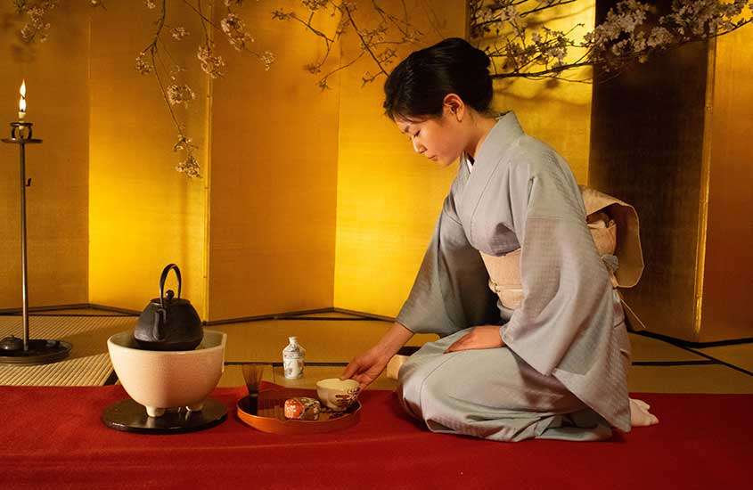 Japonesa vestindo kimono, ajoelhada em um tapete vermelho e preparando chá para uma cerimônia do chá, tradição do povo japonês