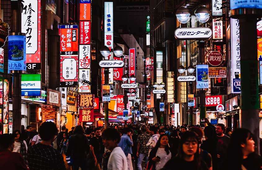 Vista noturna de uma rua movimentada em Tokyo, com várias pessoas caminhando sob uma profusão de letreiros iluminados e coloridos