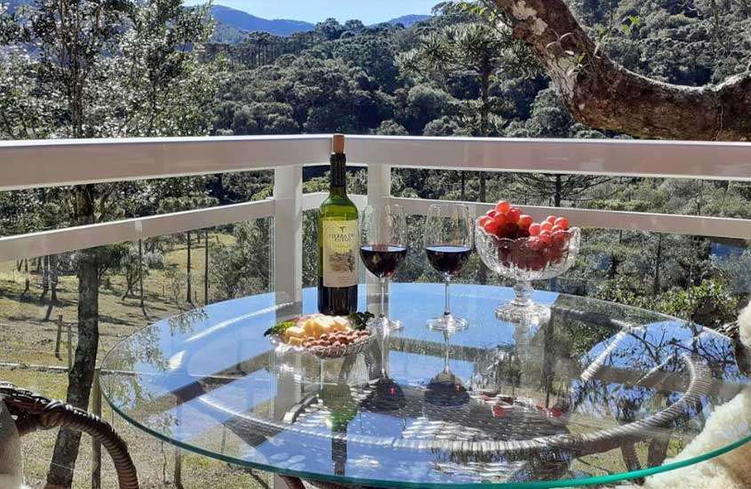 taças de vidro, uvas, frios e garrafa de vinho em cima de mesa de vidro redonda em varanda de um dos chalés em urubici para casal com vista para as montanhas