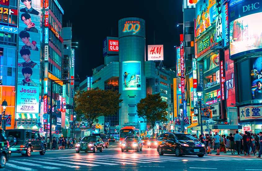 Durante a noite, uma rua movimentada de Tóquio iluminada por letreiros coloridos enquanto carros e pessoas passam