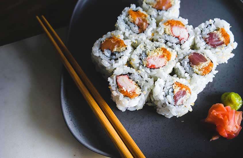 Várias peças de sushi coloridas e delicadas dispostas em um prato quadrado preto, com um par de hashis ao lado