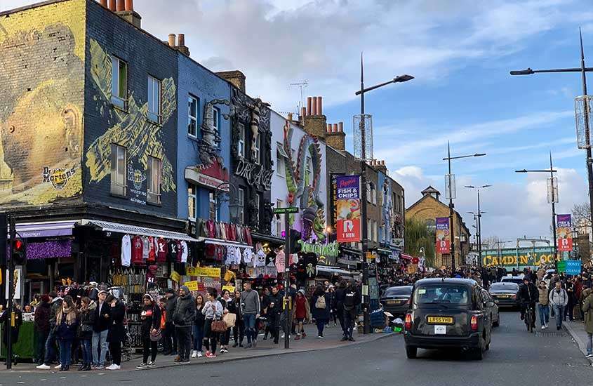 Durante o dia, carros e pedestres transitando em rua com vários outdoors coloridos em Camden Town, um lugar onde ficar em londres.