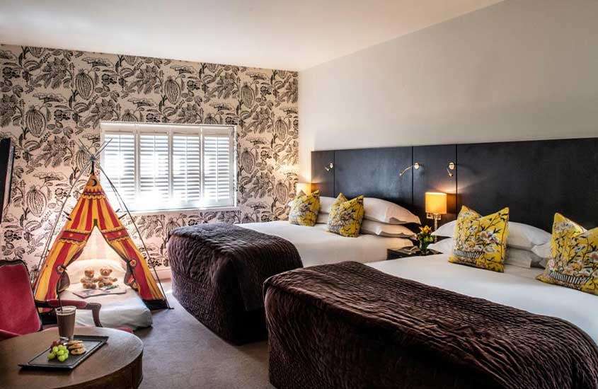 Suíte de hotel em Londres com decoração aconchegante, composta por uma mesa e cadeira de madeira, duas camas de casal e uma cabaninha infantil.