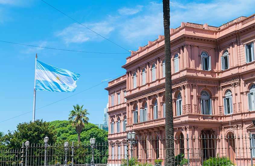 Durante um dia ensolarado, a bandeira argentina, com as cores azul e branca, hasteada em frente ao Palácio Rosa, um edifício histórico em Buenos Aires