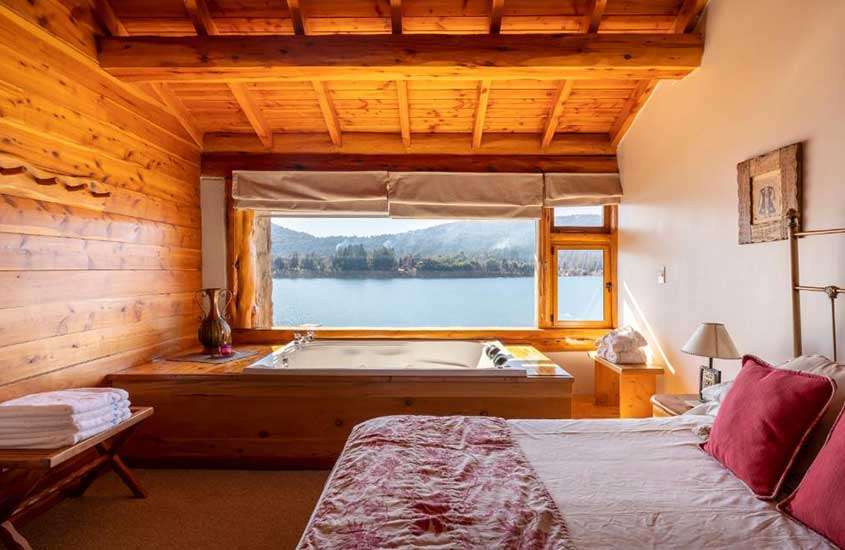 Durante o dia, um quarto de chalé em Bariloche com uma cama de casal, uma banheira de hidromassagem e uma janela com vista para um lago e montanhas