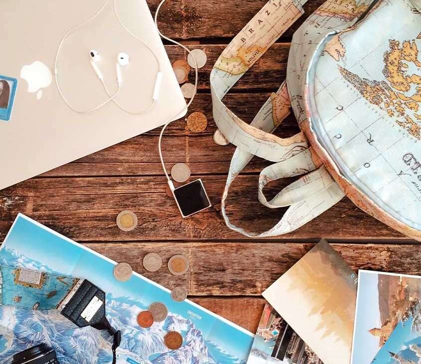 itens de viagem dispostos em cima de uma mesa de madeira, incluindo eletrônicos, mapas, moedas e uma mochila ilustrada com mapa mundi