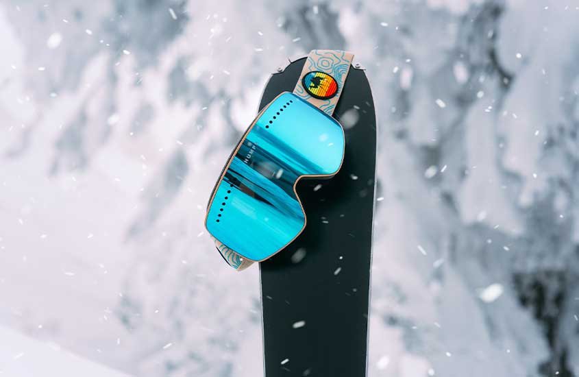 Durante o dia, óculos de proteção azuis para neve pendurados sobre uma prancha de snowboard, com uma montanha coberta de neve ao fundo