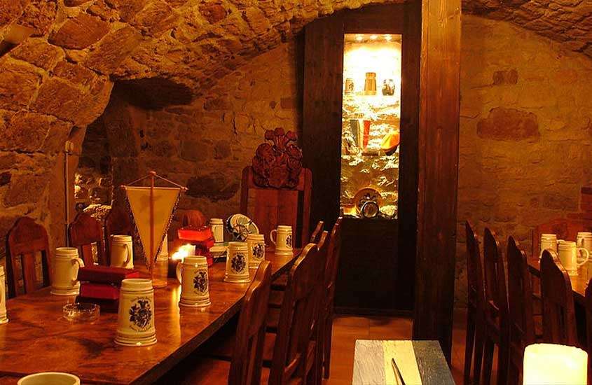 salão de restaurante rústico em Praga, que lembra taverna medieval, com canecas brancas e velas acesas em cima de mesa de madeira, rodeada por cadeiras de madeira
