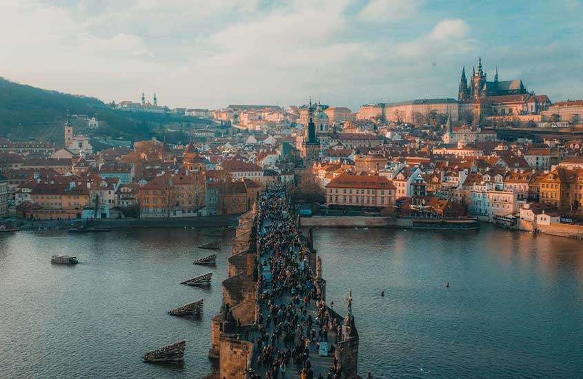 Durante o dia, vista aérea da Ponte Carlos em Praga, com pessoas caminhando sobre ela, e ao fundo, construções históricas com telhados laranjas