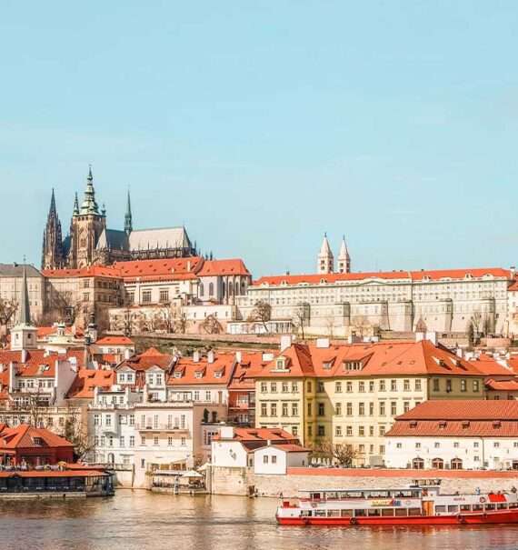 Durante um dia ensolarado, vista aérea de casas coloridas com telhado de tijolos laranja às margens de um rio em Praga, onde há um barco navegando