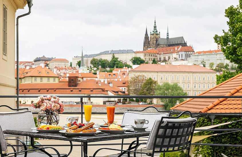 Durante o dia, cadeiras ao redor de uma mesa quadrada com diversos elementos decorativos e alimentos, incluindo vasos de flores, taças com suco, xícaras, frutas e pães, em varanda de um hotel em Praga com vista para a cidade