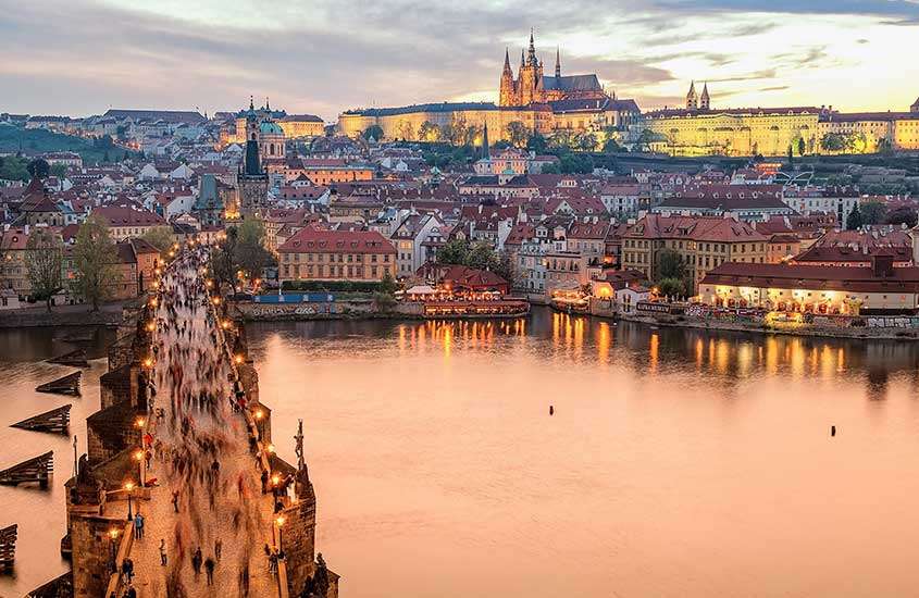 durante entardecer, vista aérea da Ponte Carlos em Praga, com pessoas caminhando sobre ela, e ao fundo, construções históricas com telhados laranjas