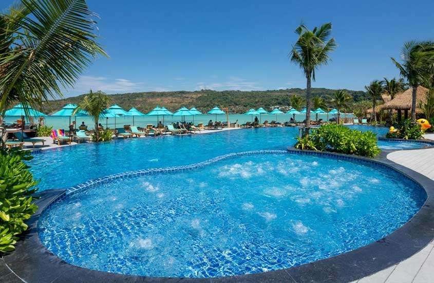 Durante um dia ensolarado, vista aérea de uma piscina ao ar livre em formato arredondado, cercada por espreguiçadeiras, guarda-sóis e árvores, em um hotel na ilha de Phi Phi, na Tailândia