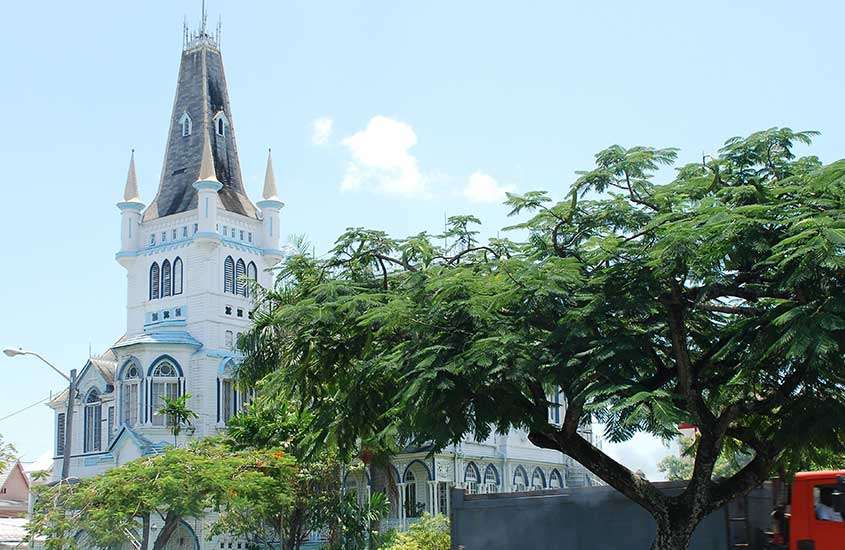 Durante um dia ensolarado, uma igreja branca e azul com telhado cinza em uma rua tranquila da Guiana, cercada por árvores.