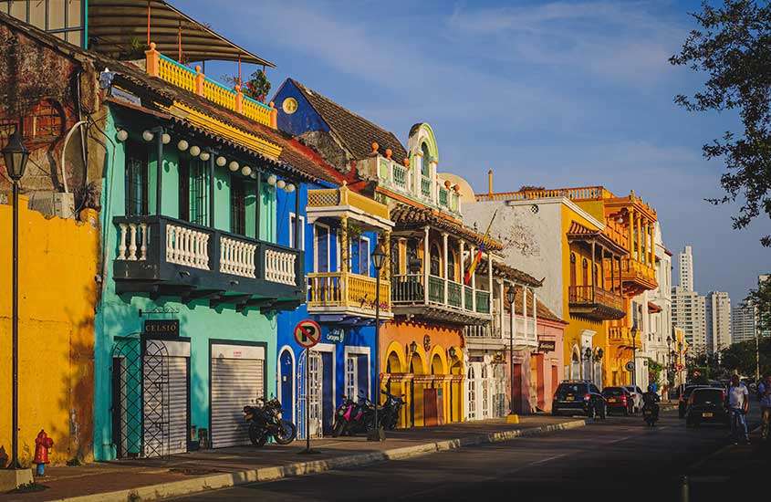 Durante o dia, carros, motos e pessoas passando em rua na Colômbia rodeada por casas coloridas.