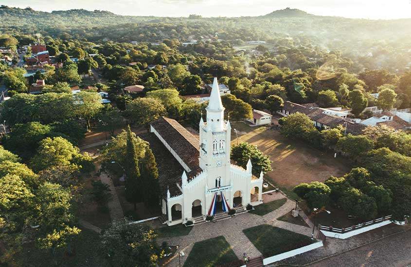 Durante o dia, vista aérea de uma igreja branca em destaque, com paisagem de árvores, casas e montanhas ao fundo no Paraguai, um dos lugares mais seguros para viajar na américa do sul.