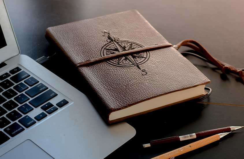Duas lapiseiras, um notebook prata aberto e um caderno com capa marrom de couro, onde há um desenho de bússola, dispostos em cima de uma mesa preta.