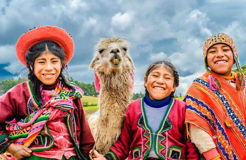 Três meninas bolivianas sorrindo e vestindo roupas coloridas, ao lado de uma lhama, durante um dia nublado.