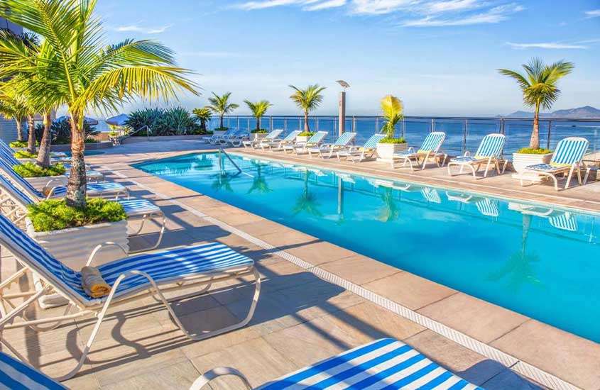 durante dia ensolarado, árvores e diversas espreguiçadeiras em frente a piscina retangular ao ar livre em hotel na barra da tijuca com vista para o mar.