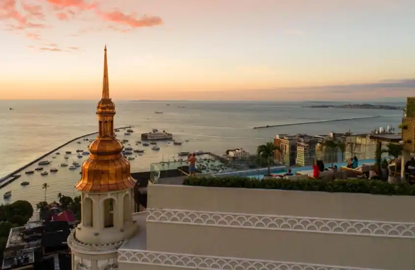 Vista aérea do Fera Palace Hotel ao pôr do sol, com a Baía de Todos os Santos ao fundo e céu colorido em tons de laranja e rosa