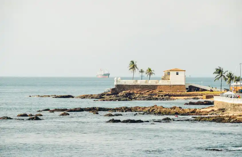 Fortaleza em Salvador, Bahia, com coqueiros à beira-mar e um navio ao fundo durante o dia